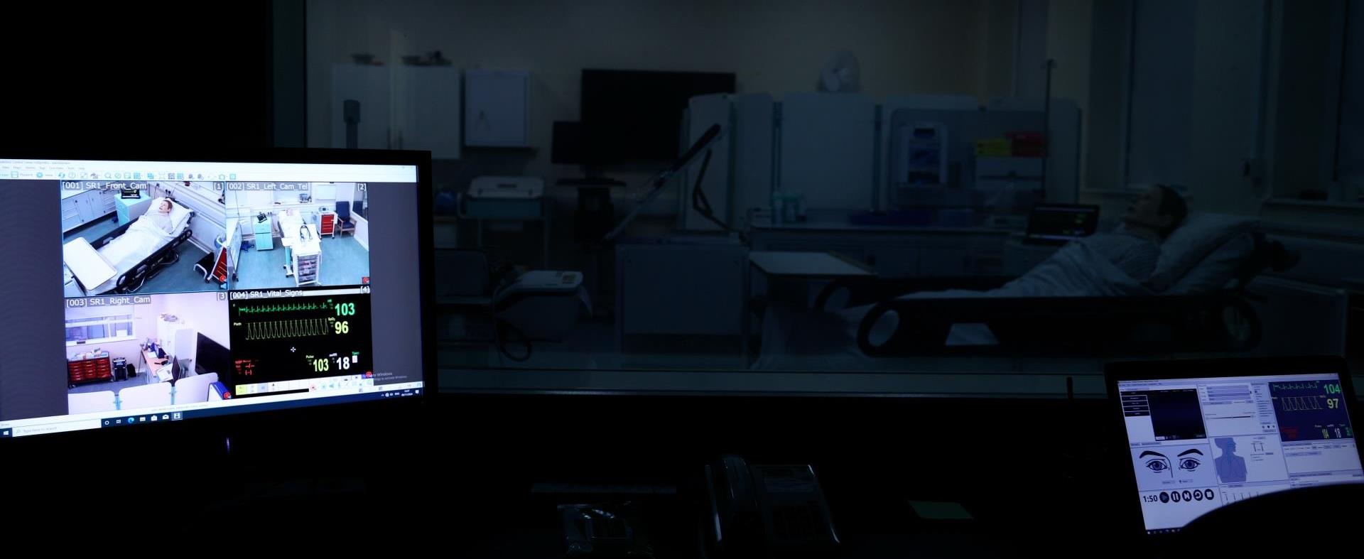 A control room with monitors showing video feeds and vital signs from a clinical simulation session. A mannequin is visible through the observation window.”