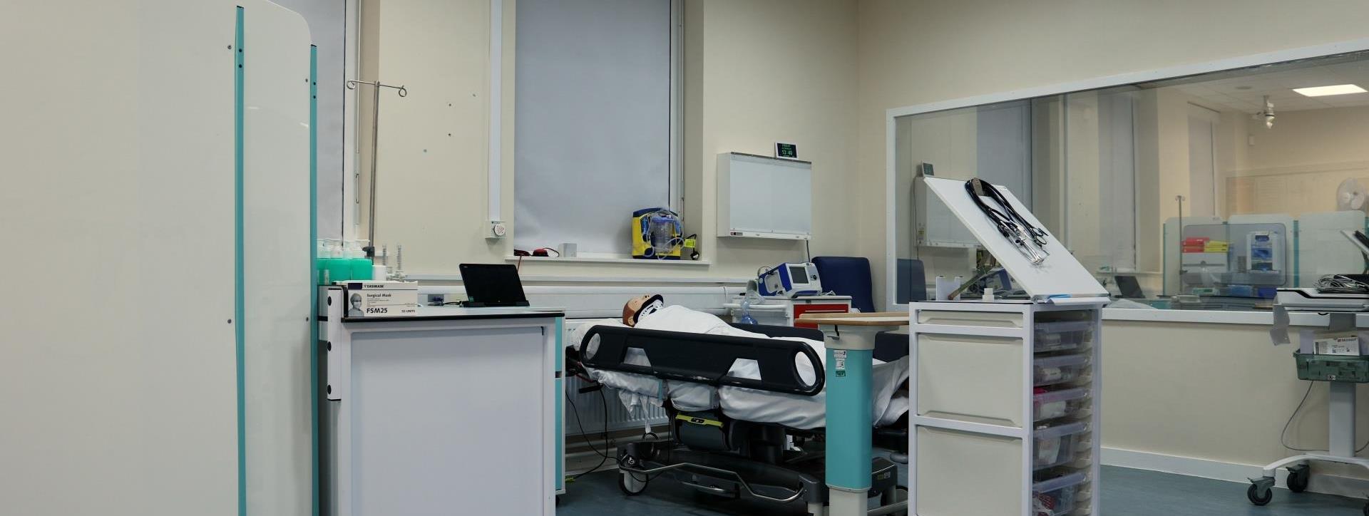 “A clinical simulation room featuring a hospital bed with a mannequin, a rolling cart with medical supplies, and a monitor. The room has a clear glass observation window.”