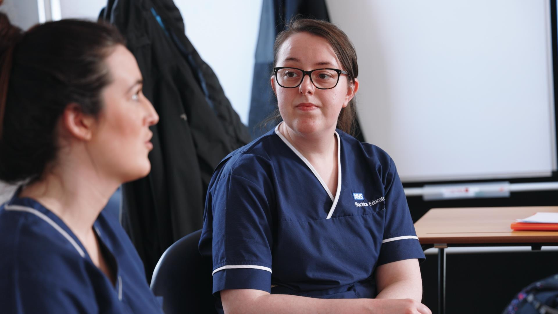 Two healthcare professionals in blue NHS practice education uniforms are seated and engaged in a discussion. One person is looking towards the other, listening intently, with a whiteboard and various teaching materials visible in the background