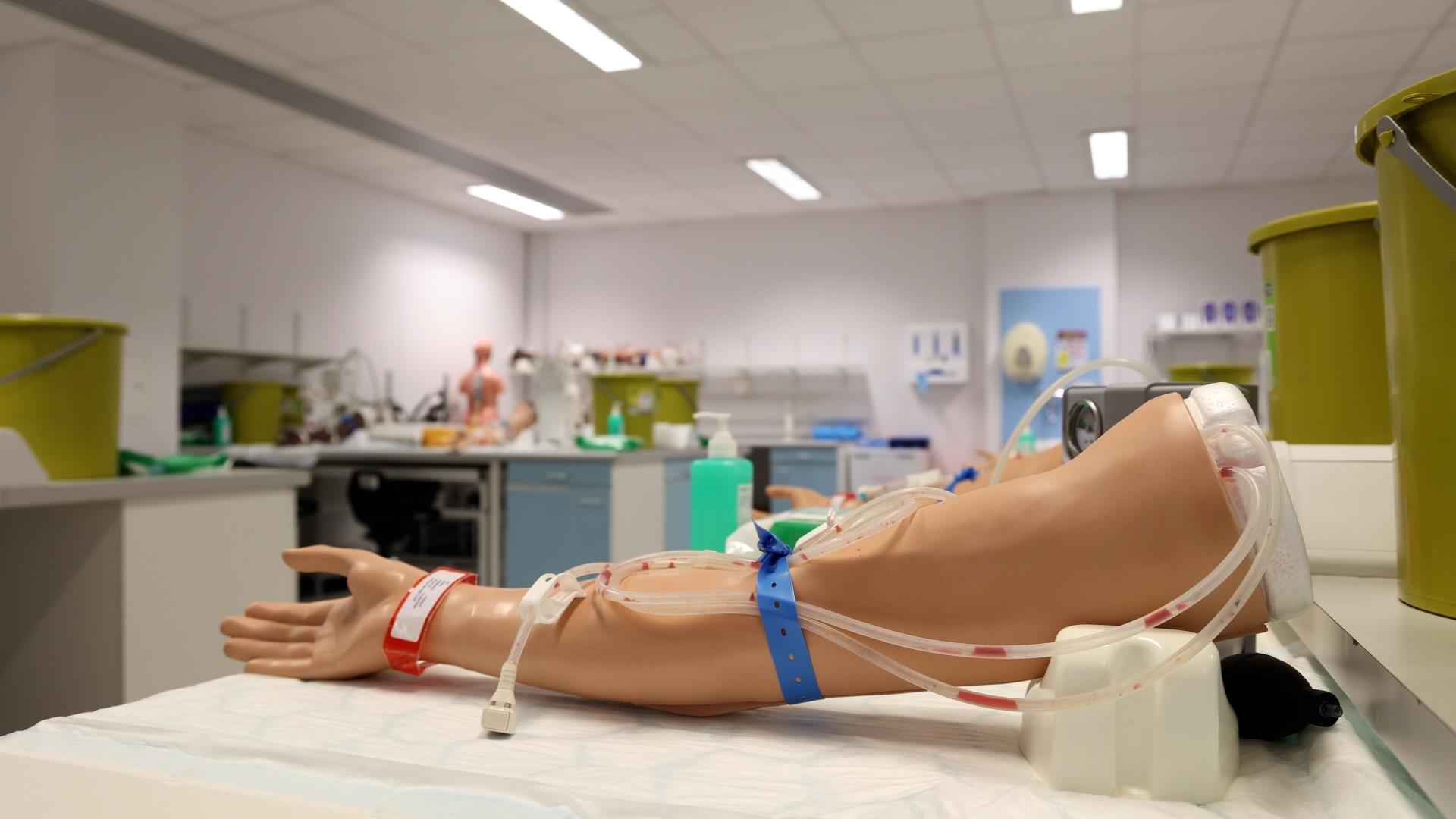 “Close-up of a medical training arm with intravenous lines, blood flow simulation, and a blood pressure cuff. The lab setting includes equipment and mannequins in the background.”