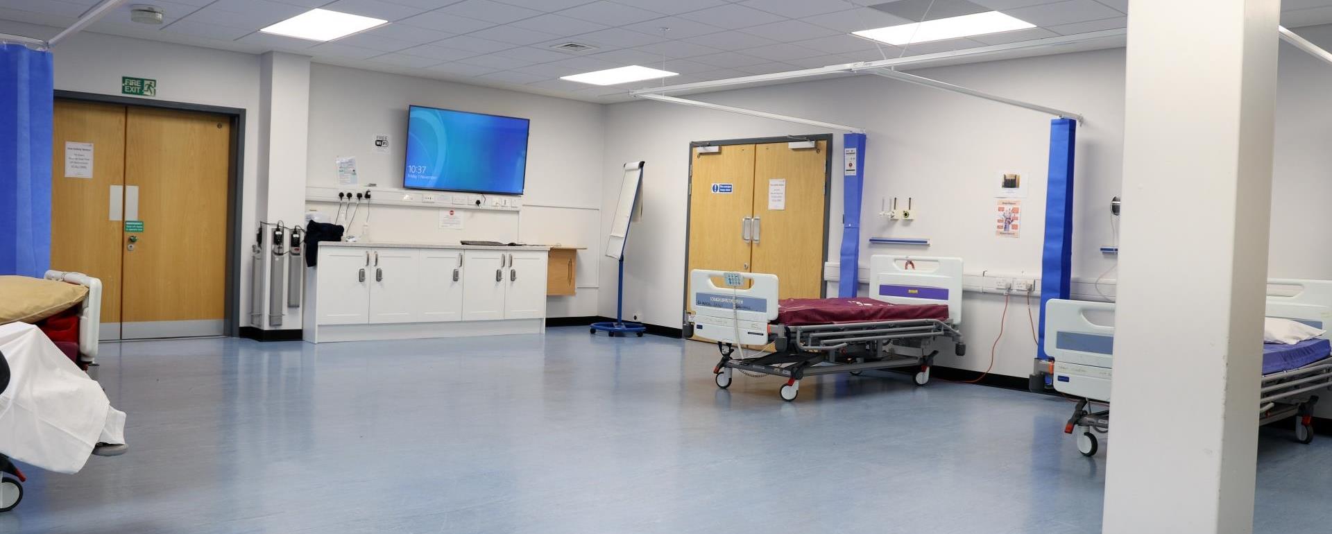 “A hospital ward simulation room with empty patient beds, medical equipment, and a TV screen on the wall. The space includes white cabinets and an open area for training.”